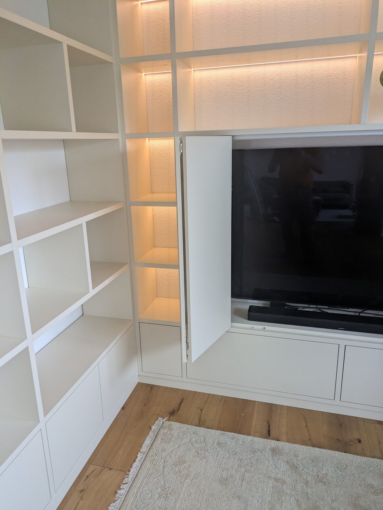 Weißer Einbauschrank vom Boden bis zur Decke mit offenen Fächern, indirekt beleuchteten Rückwänden und einem Fernseher hinter geöffneten Falttüren.